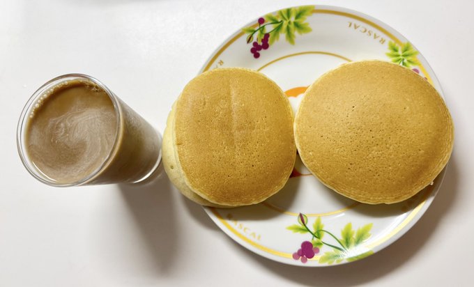 A glass of milky coffee sits next to a plate decorated with grape illustrations and the word 'MACAU', holding two fluffy golden pancakes.