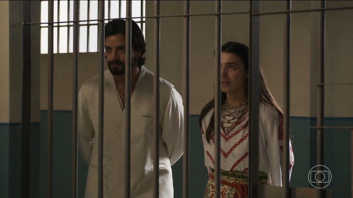 A man with a beard wearing a white vest over a white shirt and a woman in a patterned dress stand side by side behind prison cell bars in what appears to be a television drama scene.