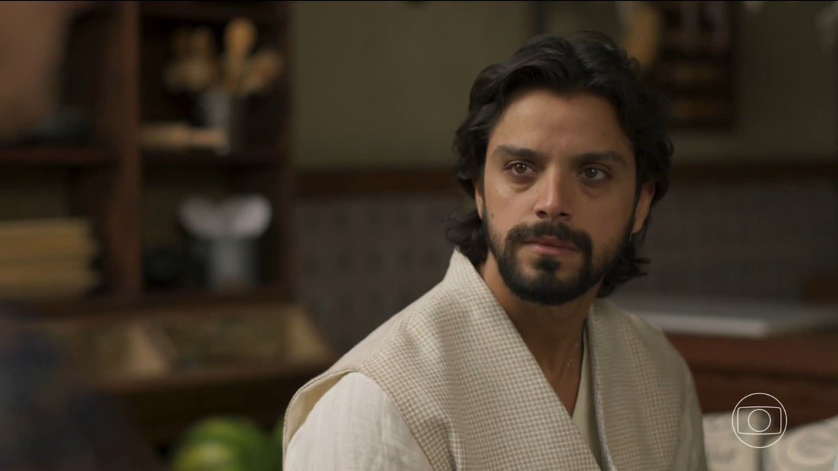 A dark-haired, bearded man wearing a checkered vest over a white shirt gazes thoughtfully off-camera, set against a warmly lit interior with shelves in the background, with the Globo network logo visible in the bottom right corner—likely capturing a lighthearted moment from the Brazilian telenovela referenced by the #ANobrezaDoAmor hashtag.