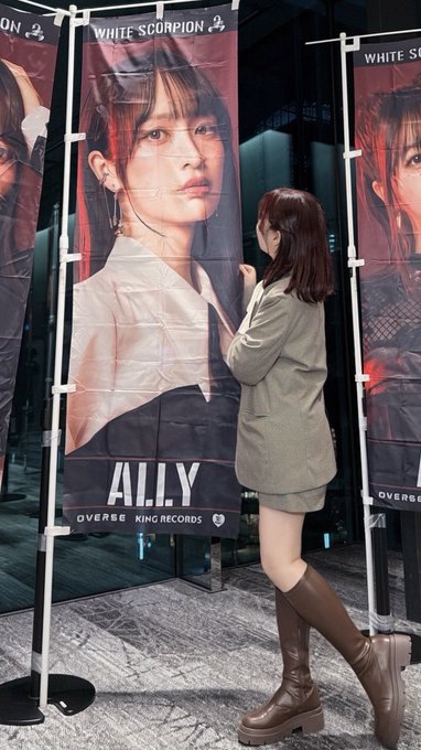 A woman in a green blazer and knee-high boots stands next to a large promotional banner for 'ALLY' from 'WHITE SCORPION' on 'OVERBSE KING RECORDS'.