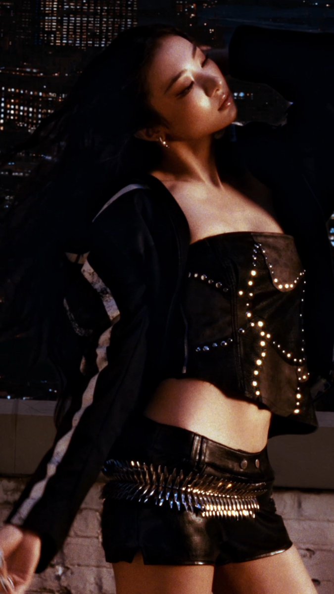 A woman with long dark hair poses on a rooftop at night wearing a studded black leather corset and matching shorts with a spiked belt.