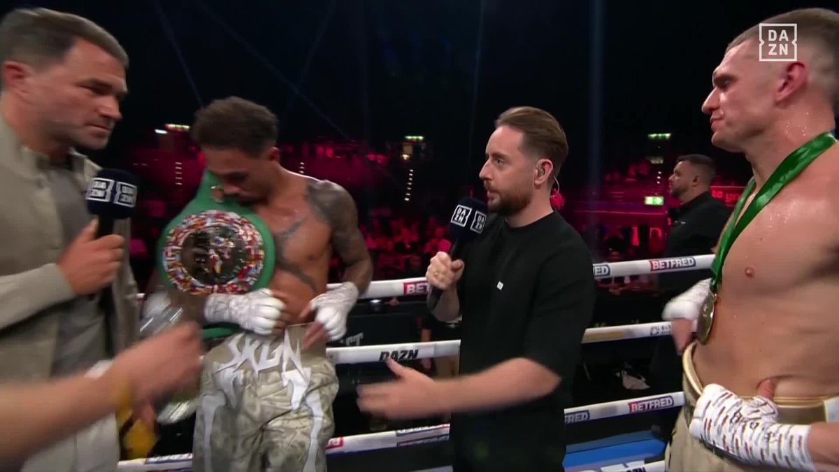 Boxing promoter Eddie Hearn (center) conducts a post-fight interview in the ring with two shirtless fighters wearing championship belts and medals, with "DAZN" and "BETFRED" branding visible on the ring ropes and microphones, capturing the post-main event moment following the Whittaker vs. Wallace bout.