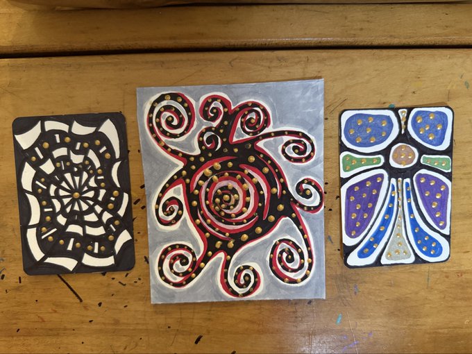 Three hand-drawn abstract artworks laid out on a wooden surface: a black and white mandala pattern on the left, a swirling red, black, and gold octopus-like design in the center, and a blue and purple dotted figure on the right.