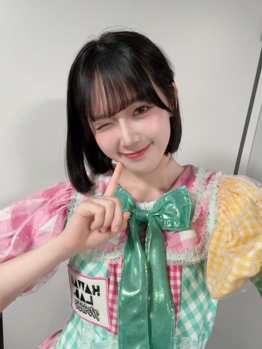A woman winks and points upward while wearing a colorful outfit featuring a large green bow and a 'HAPPY HALLOWEEN' name tag.