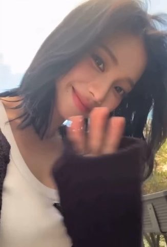A young woman with shoulder-length dark hair smiles softly at the camera while wearing a white top and dark cardigan, with her hand raised near her face in an outdoor setting.