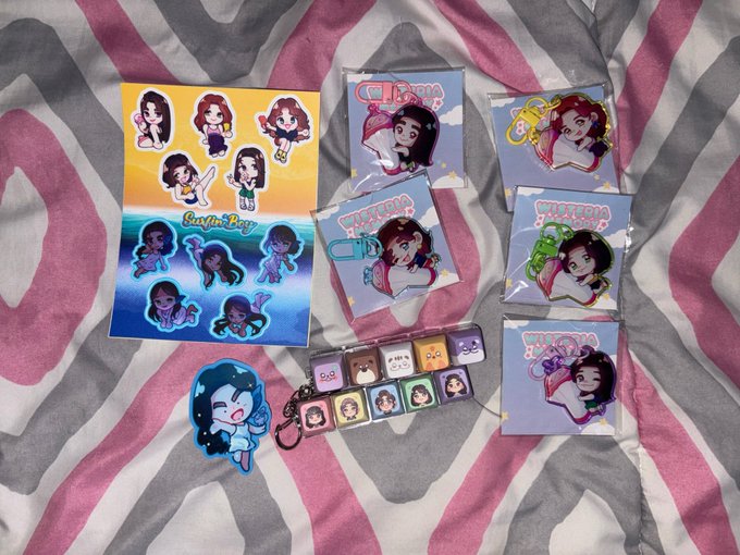 A collection of colorful anime-style stickers and merchandise featuring various character designs are arranged on a pink and gray zebra-striped fabric surface.