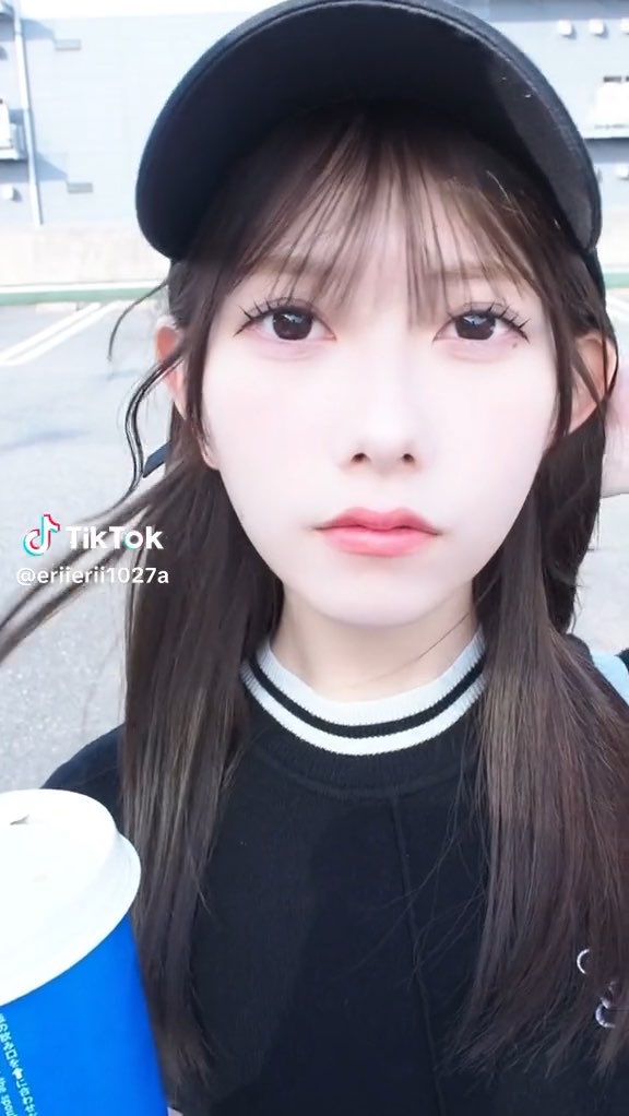 A TikTok selfie video of a young woman wearing a black cap and black sweater with white stripes, with the TikTok handle '@eririri1027a' visible.