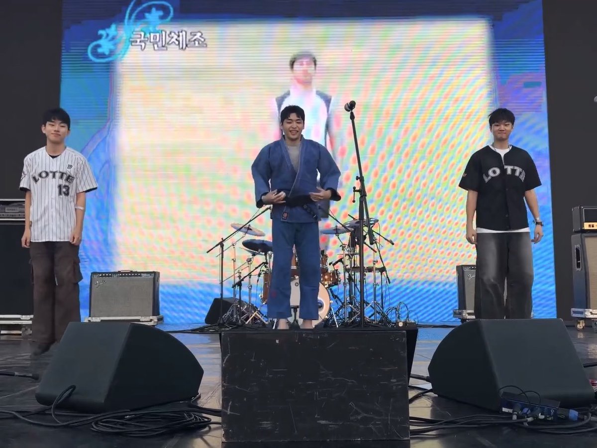 Three young men stand on a stage with a Korean banner reading '국민체조' (Gungmin Chumcha) on the screen behind them, with a performer visible on the large display.