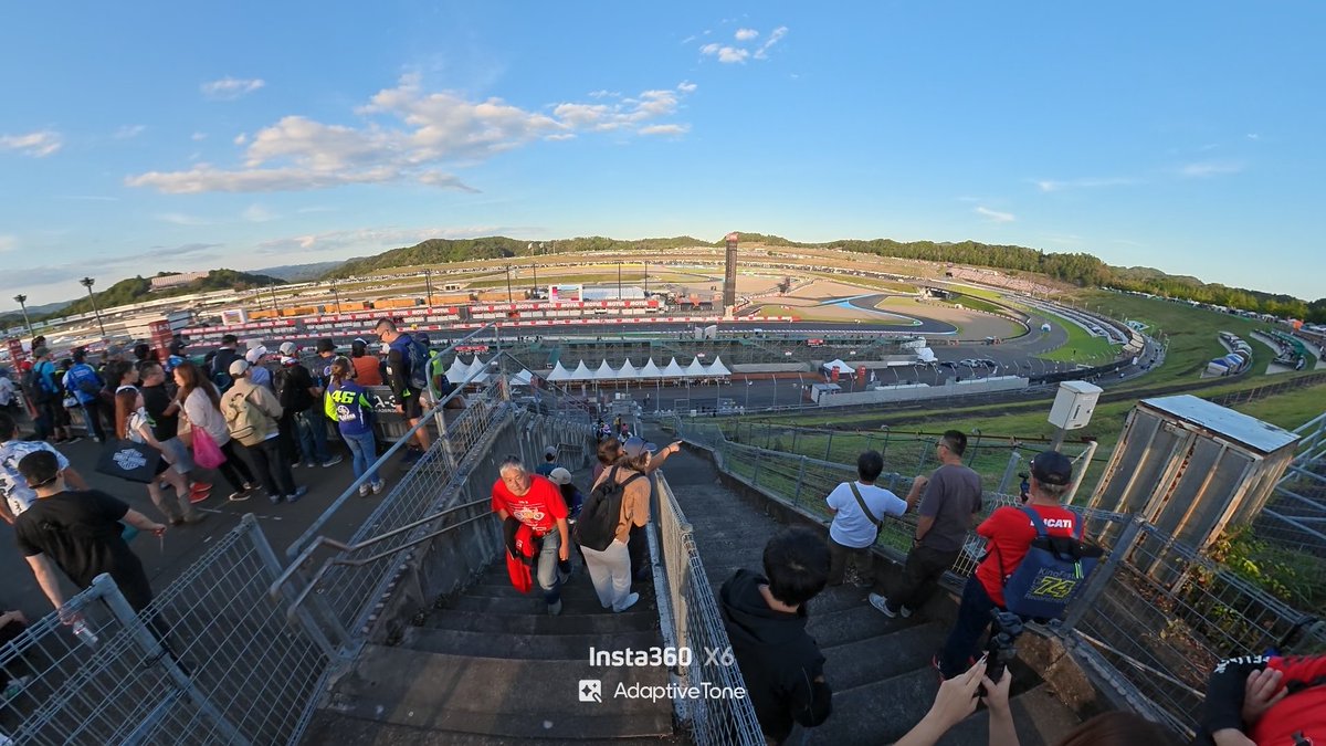 A crowd of spectators walking down concrete stairs toward a sprawling MotoGP-style racing circuit, captured with an Insta360 X6 camera.