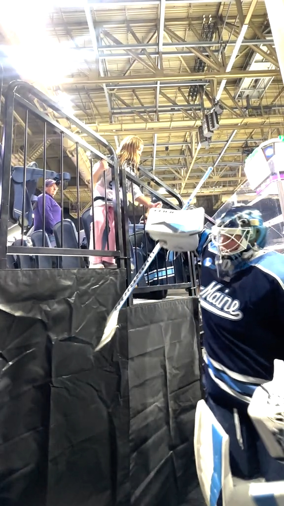 A hockey player in full gear, wearing a dark blue 'Maine' jersey and mask, stands near the rink boards where fans are seated above.