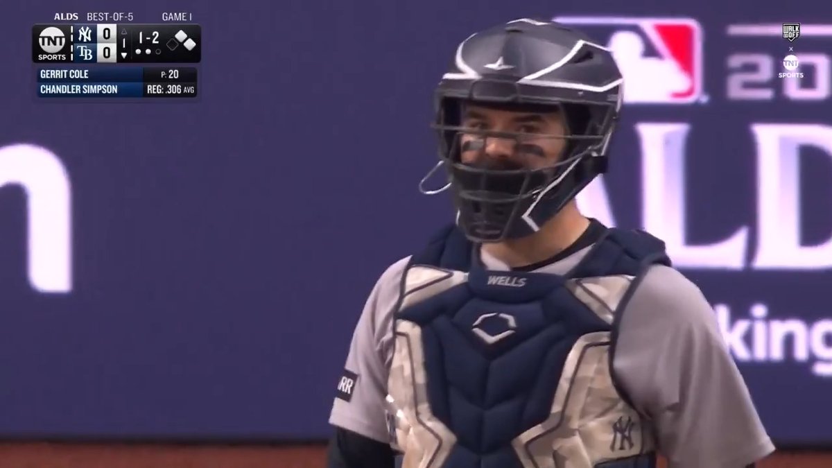 Yankers catcher Austin Wells smiles behind his catcher's mask during Game 1 of the ALDS against the Rays, with the score tied 0-0 in the top of the first inning with a 1-2 count on batter Chandler Simpson against Gerrit Cole. The image captures Wells's confident demeanor following his successful challenge call, helping Cole navigate a tough at-bat against Tampa Bay.
