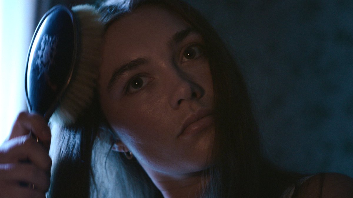A woman with dark hair holds a hand mirror up to her face, gazing intently into it with wide, focused eyes in dramatic blue lighting. The image captures the caption's sentiment perfectly—the intensity in her expression mirrors the feeling of someone wishing Florence Pugh could narrate the private, introspective moments of their life.