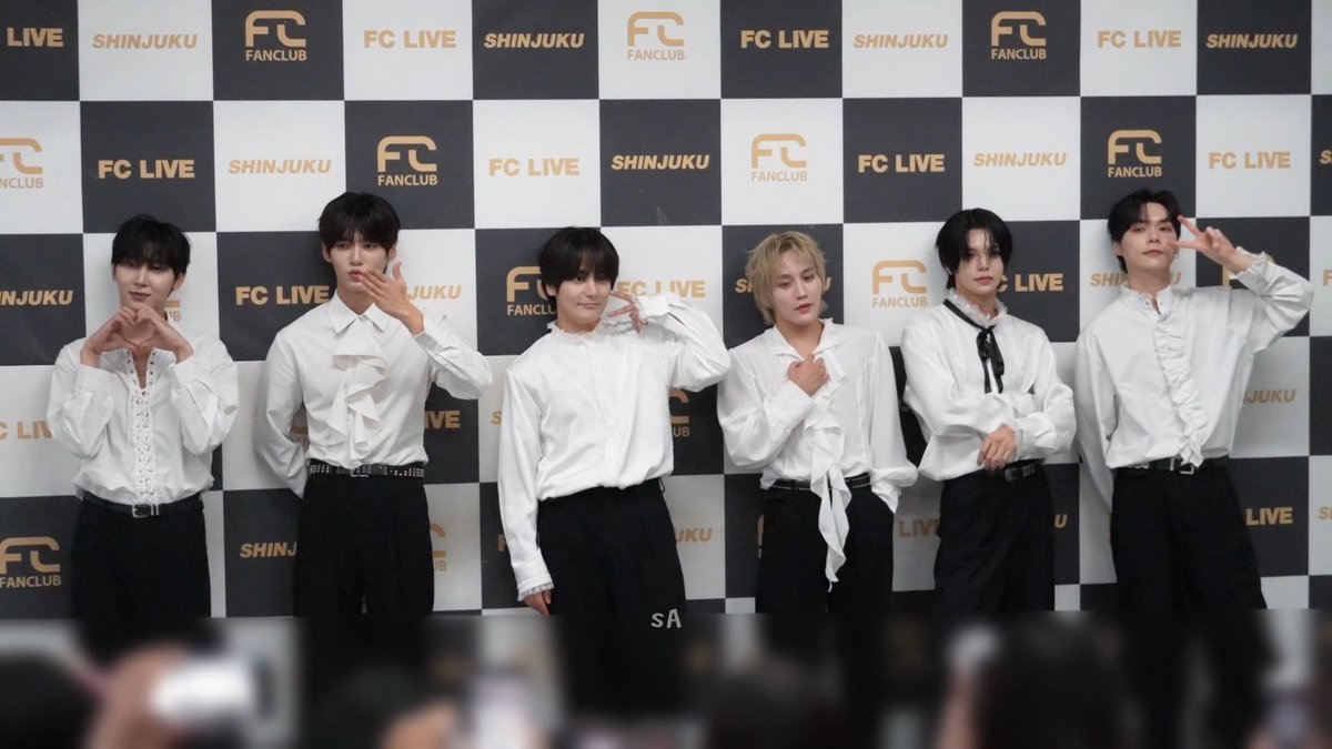 Six members of a K-pop group pose in white shirts and black pants in front of a black and gold checkered backdrop with 'FC LIVE', 'SHINJUKU', and 'FC FANCLUB' branding.