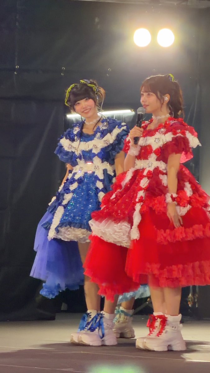 Two young women on stage wearing elaborate, frilly idol costumes—one in blue and white, one in red and white with white platform boots—with the red-costumed member speaking into a microphone.