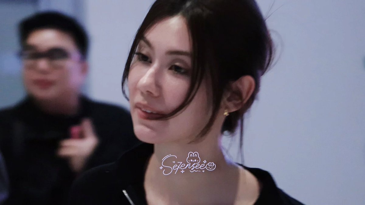 A close-up shot features a young woman with dark hair in an updo, wearing a black jacket, with the watermark "Se7enBae" and a bunny logo visible. In the blurred background, a man in sunglasses appears to be clapping, consistent with the post's references to CHANEL Spring Summer and Paris Fashion Week, likely capturing a moment from the CHANEL SS27 show featuring Lena Miu.