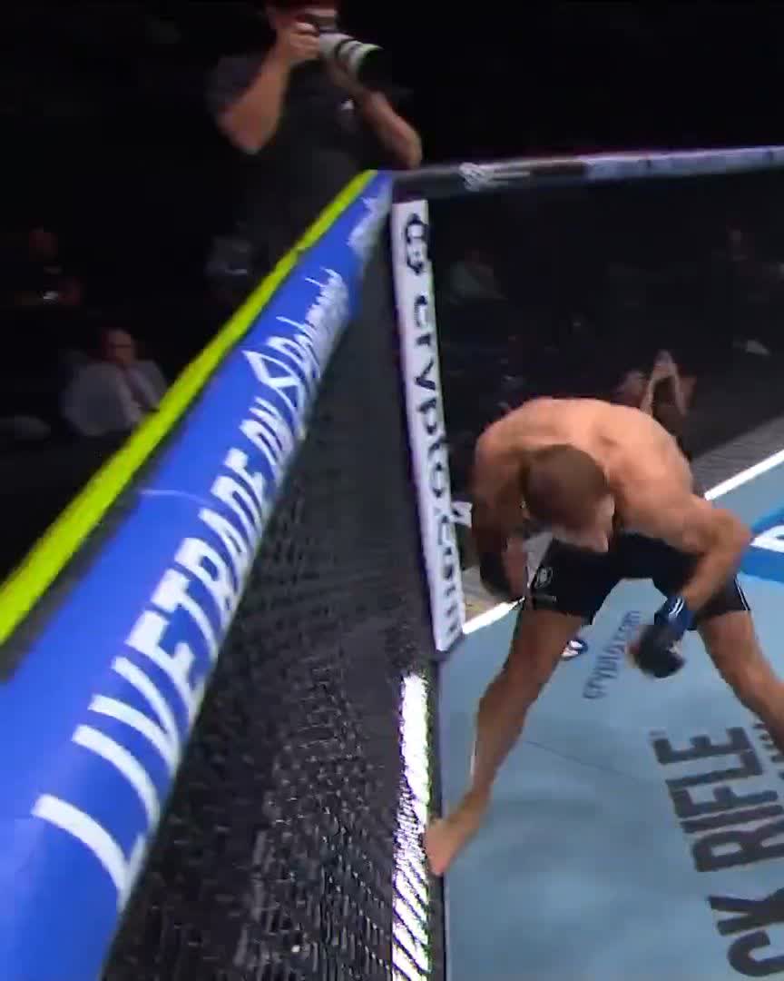 A shirtless MMA fighter in a UFC octagon leaning over the cage padding, appearing to recover or tend to himself after a fight, with a cameraman visible in the background.