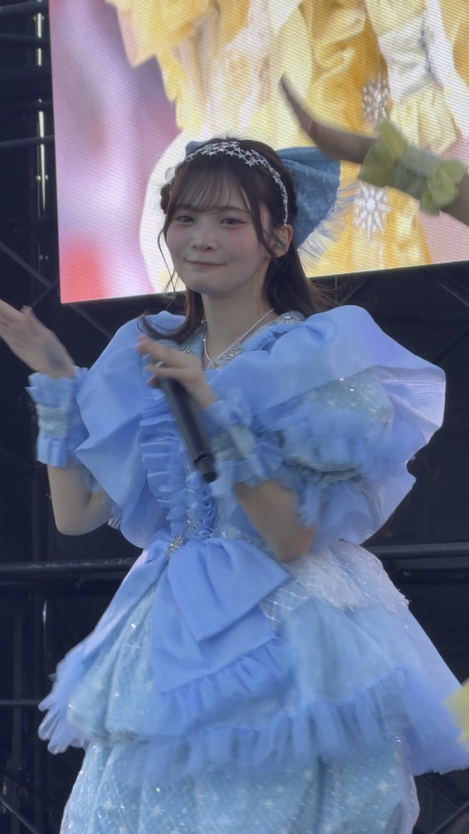 A young woman performs on stage wearing an elaborate blue princess-style dress with puffy shoulders and a matching bow headband, holding a microphone in front of a large screen.