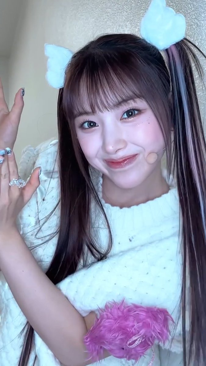 A young woman with long brown hair and pigtails poses for a selfie, wearing a white knit top, cat ear hair clips, a pink fluffy wrist accessory, and a ring on her finger.