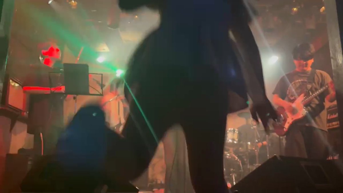 A silhouetted performer stands dramatically in the foreground of a dimly lit live music venue, with a guitarist playing on stage behind them under green and red stage lights. The intense, immersive concert atmosphere—complete with smoke and close-up staging—matches the post's teasing caption, suggesting the scene captures the overwhelming experience of someone attending their first live house show.