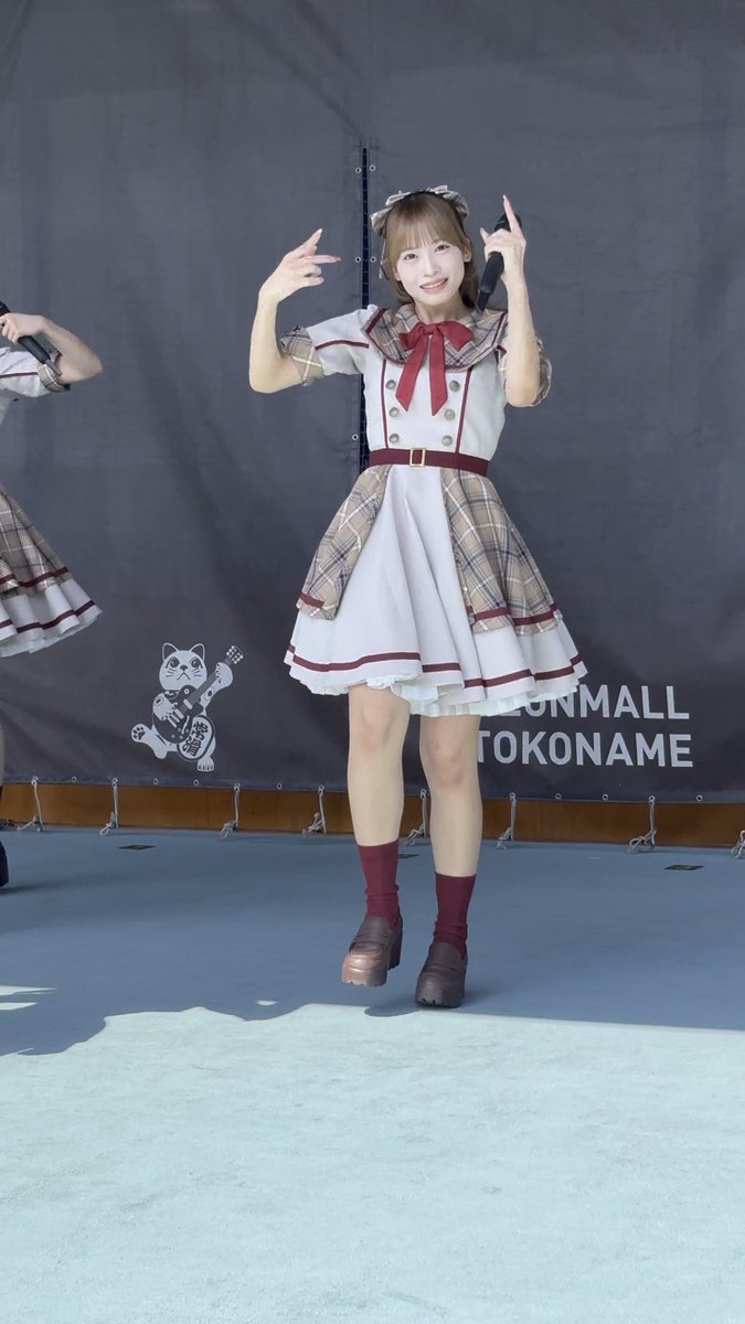 A young woman in a white and plaid dress with red accents performing on stage, making peace signs with both hands, with 'AEON MALL TOKONAME' visible on the backdrop behind her.
