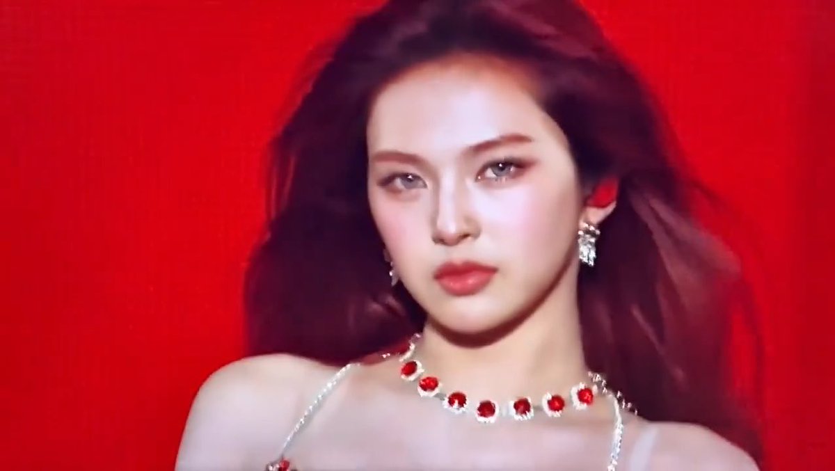 A glamorous woman with dark hair, wearing diamond and ruby jewelry, looks directly at the camera against a solid red background.