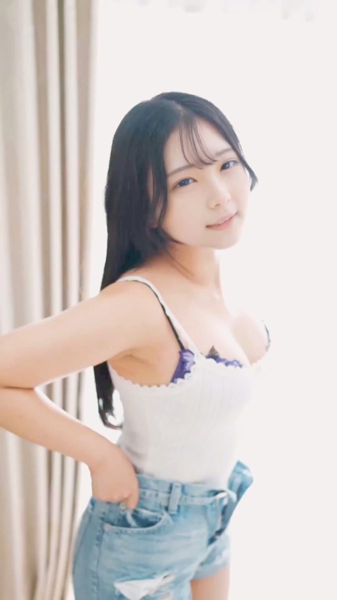 A young woman with long dark hair poses in a white camisole top and light blue denim shorts in a brightly lit setting with white curtains.