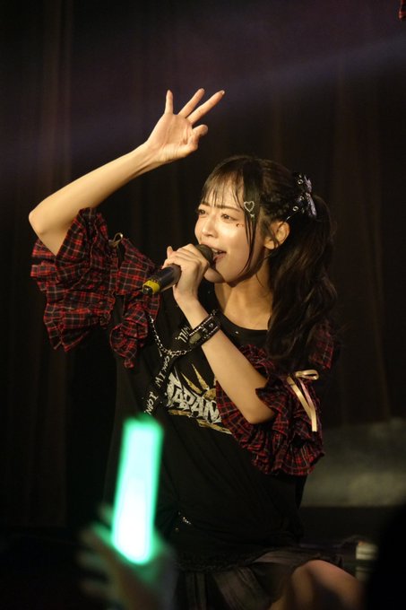 A young female performer with dark hair sings into a microphone on stage, wearing a black outfit with red plaid details and making a peace sign with her hand.