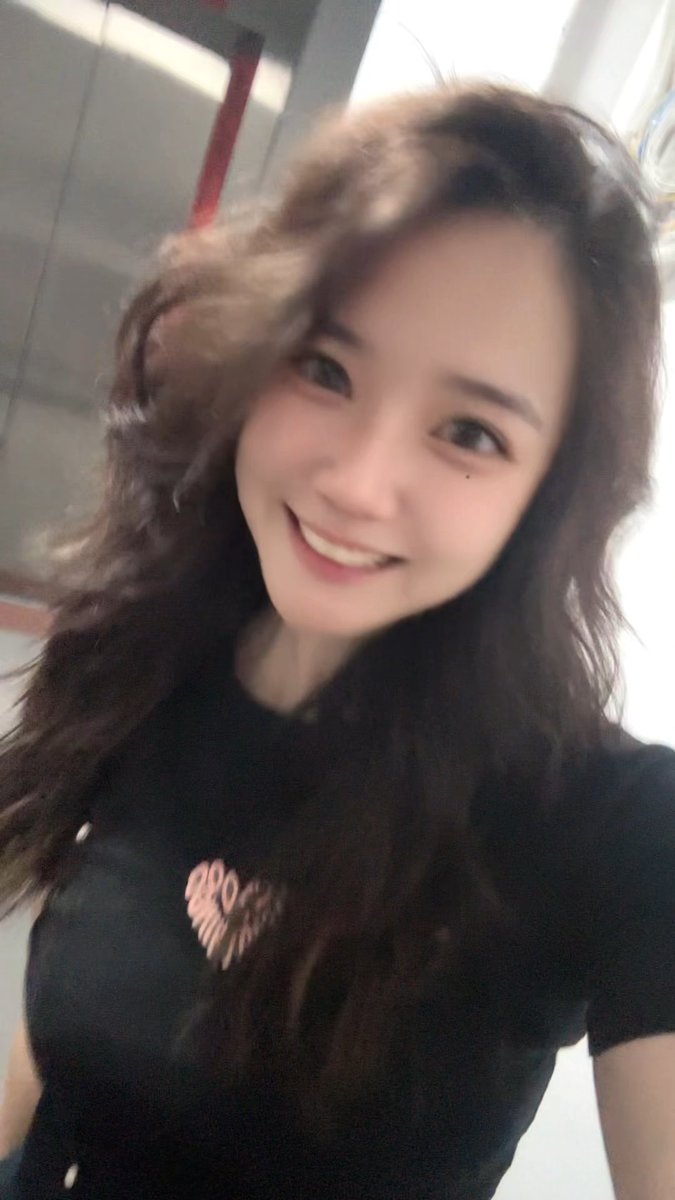 A selfie of a smiling woman with long, wavy brown hair wearing a black t-shirt with pink lettering.