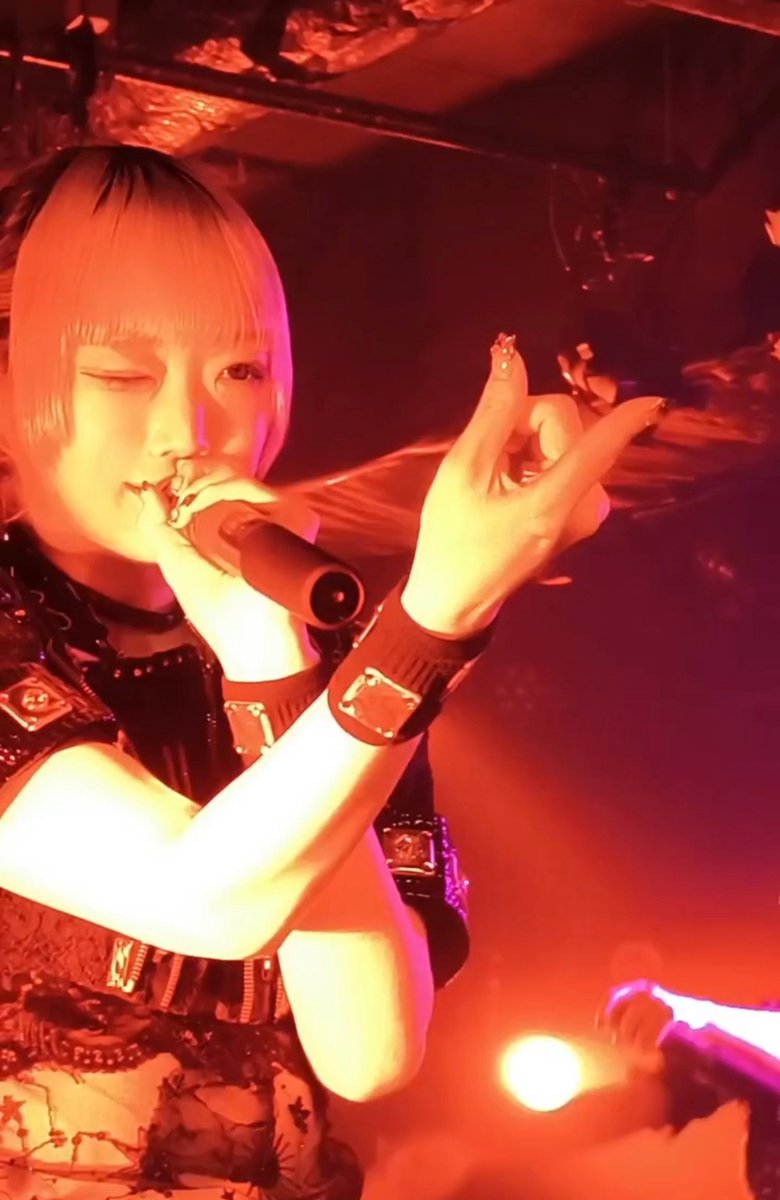 A performer with short blonde hair and bangs singing into a microphone on a stage bathed in warm red and orange lighting, wearing a dark outfit with studded wristbands and long decorated fingernails.