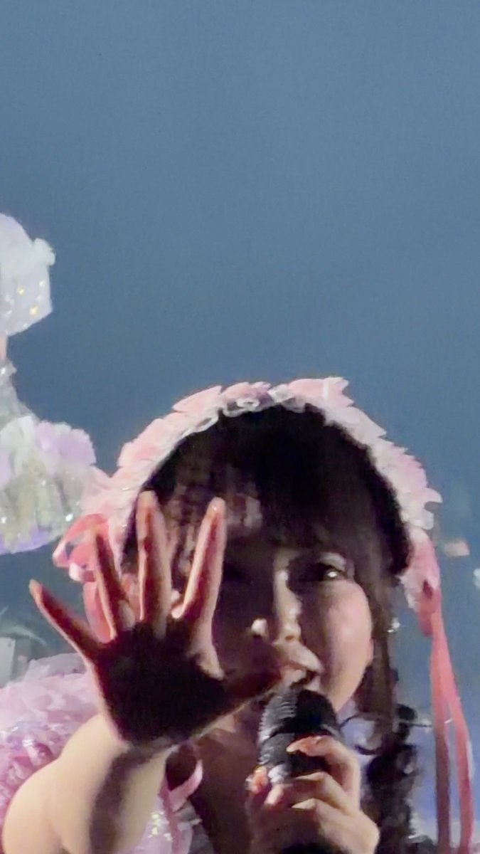 A young woman with dark hair, wearing a pink frilly headpiece, holds her hand up toward the camera while singing into a microphone against a blue background.