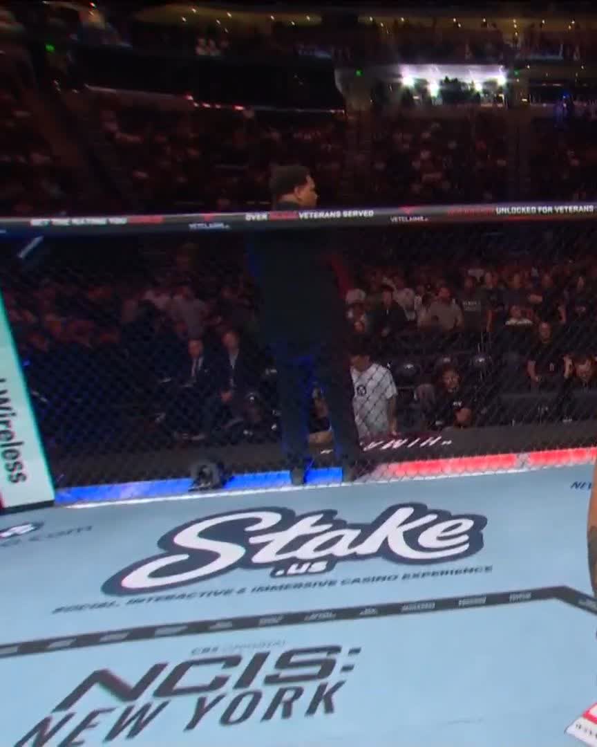 Inside a UFC octagon, a referee in black walks across the canvas displaying 'Stake.us', 'NCIS: NEW YORK', and 'VETERANS SERVED' sponsor logos while the arena crowd looks on.