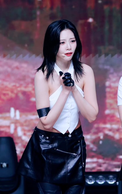 A young woman with black hair wearing a white halter top, black leather skirt, and black gloves performs on stage with her hands clasped together.