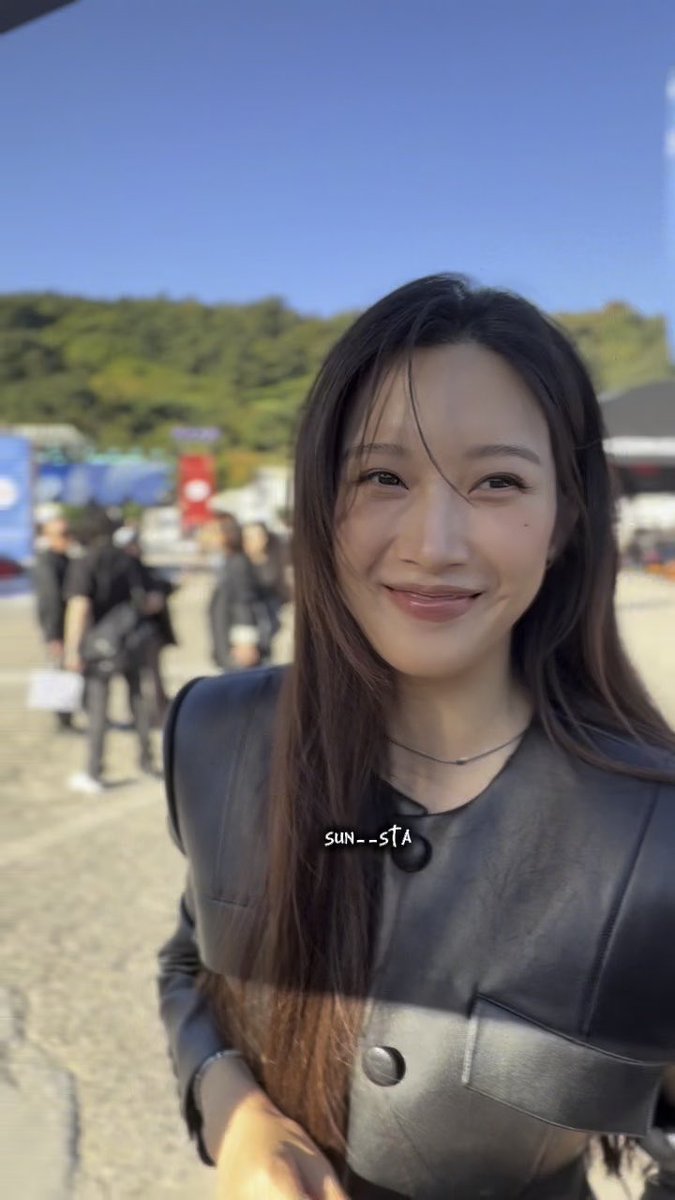 A selfie of a smiling young woman with long dark hair wearing a black leather jacket outdoors, with a blue sky and hills in the background, watermarked 'SUN--STA'.