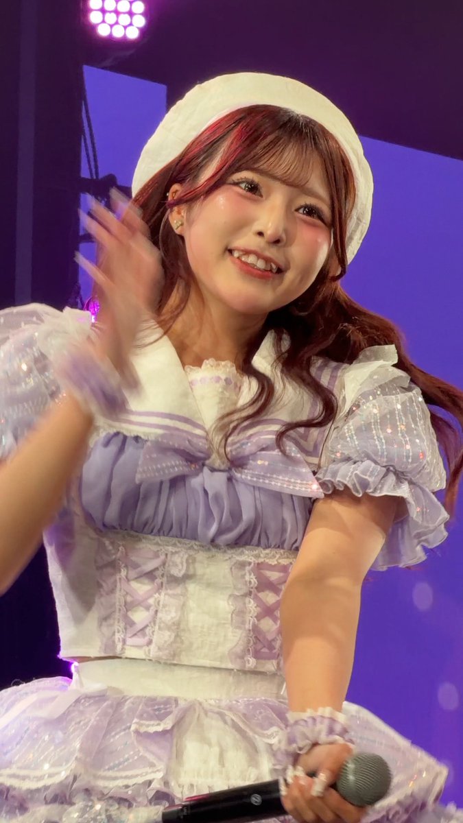 A young woman smiles on stage, wearing a frilly white and light purple dress with a matching beret, and holds a microphone in her hand.