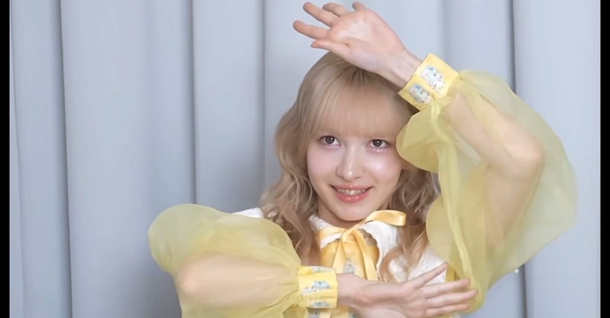 A young woman with blonde curly hair wearing a yellow and white outfit with puffy sleeves, posing with one hand raised above her head and the other near her waist against a gray curtain backdrop.