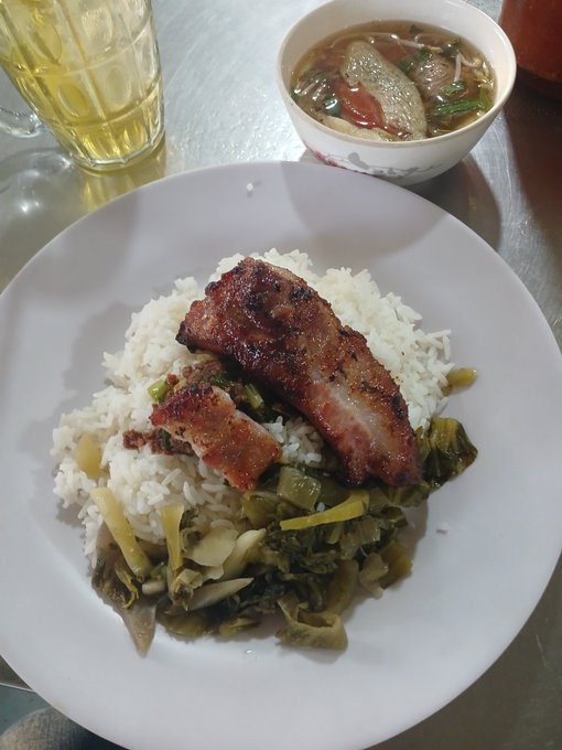 A plate of white rice topped with grilled meat and pickled greens is served alongside a bowl of soup, with a glass of yellow beverage nearby.
