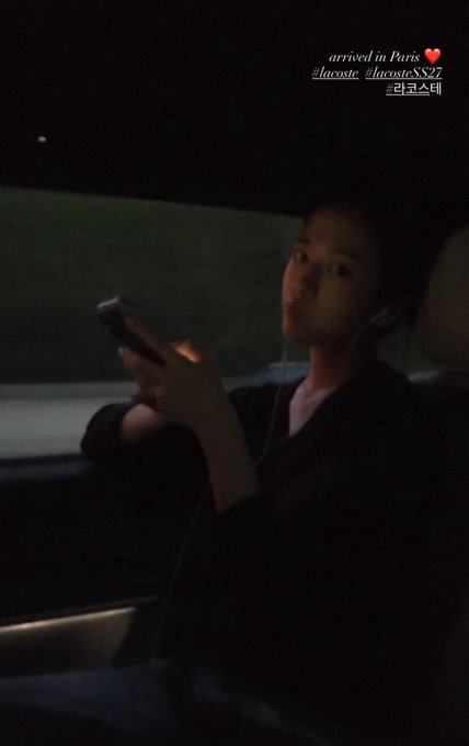A young woman sits in a dark car using a smartphone while wearing earphones, with on-screen text reading 'arrived in Paris #lacosta #lacostaSS27' and a Korean tag.