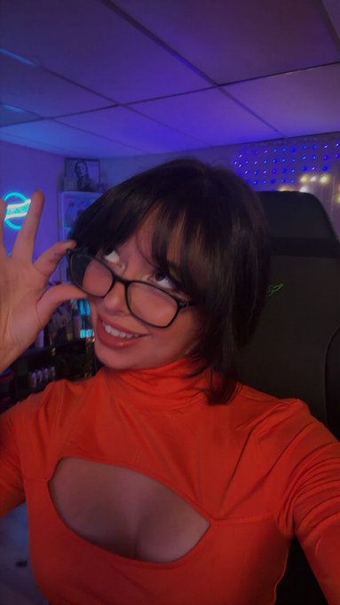 A young woman with short dark hair, glasses, and an orange top making an OK hand gesture in a room with neon lights and LED panels in the background.