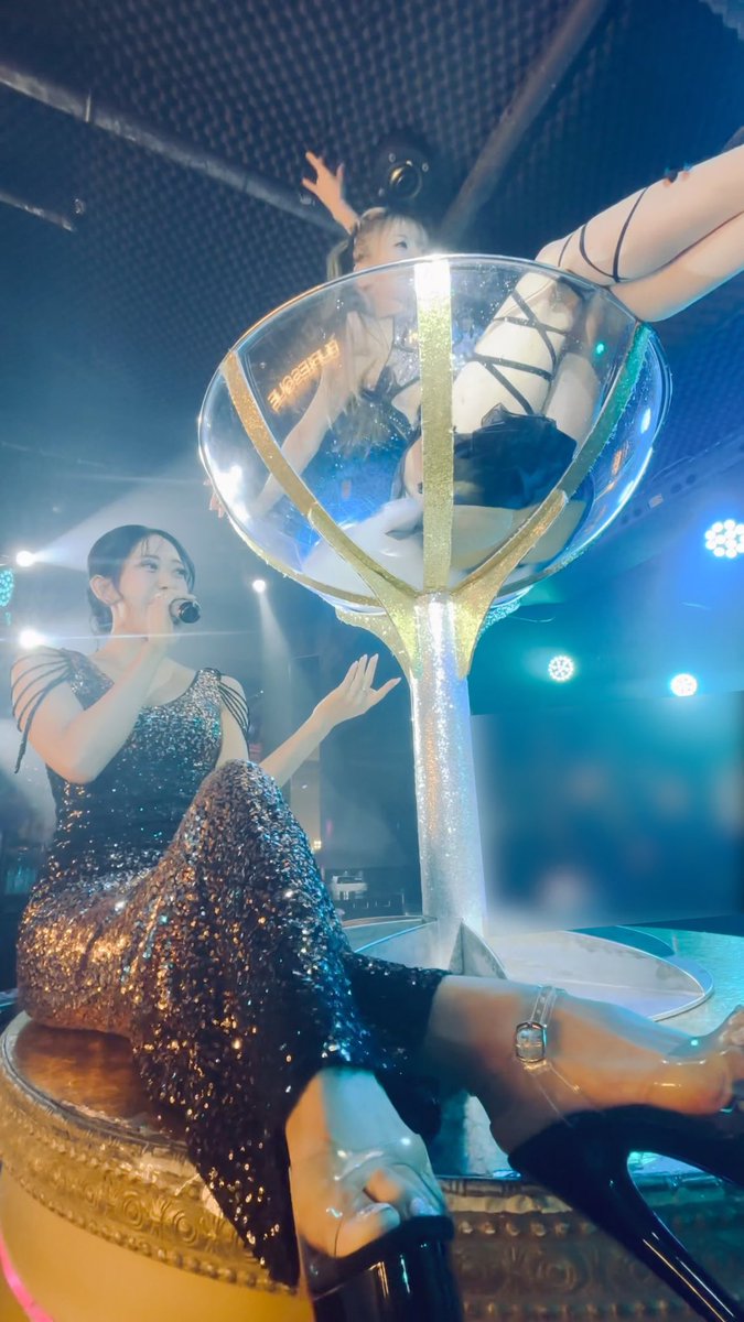 A performer in a sparkly sequined gown sitting on the base of a giant champagne glass prop during a concert, while another performer poses inside the glass above.