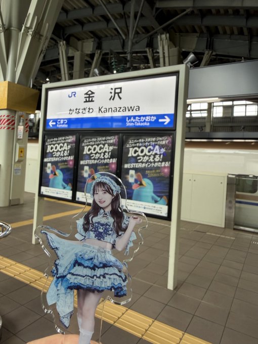 A cardboard cutout of a young woman in a blue frilly dress stands in front of a JR Kanazawa station sign with ICOCA advertisements visible in the background.