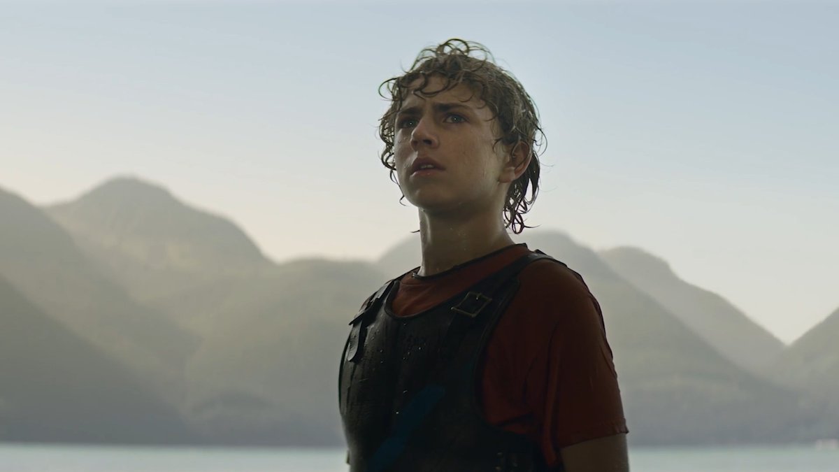 A young person with wet, curly hair and a red shirt under dark armor stands against a backdrop of mountains and water, gazing upward with a contemplative expression. The image, appearing to be from Percy Jackson and the Olympians on Disney+, illustrates the post's point that Disney's strong social media presence significantly boosts anticipation for the show's third season, unlike Netflix's weaker promotional reach.