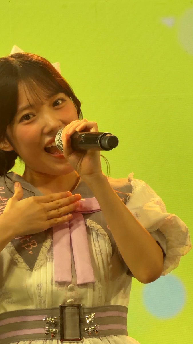 A young woman with brown hair, wearing a white and gray dress with a pink ribbon, smiles brightly while singing into a microphone against a bright green background.