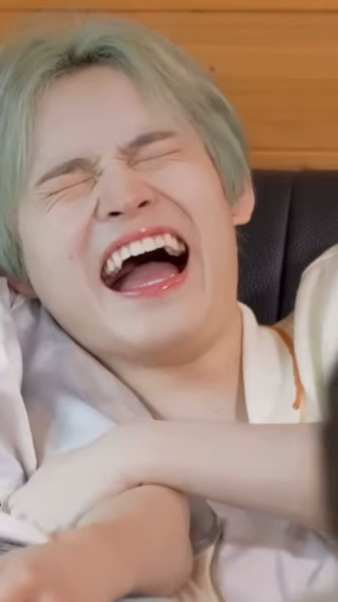 A young man with light green hair is shown laughing hysterically with his eyes closed and mouth wide open.