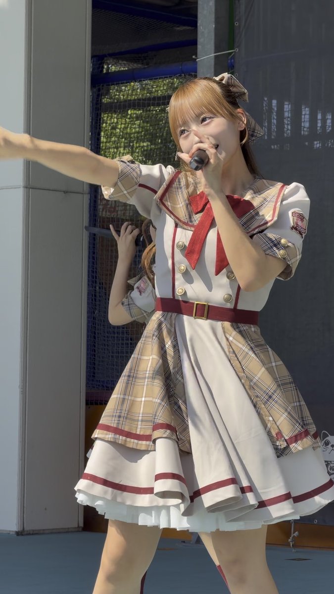 A performer on stage wears a white, beige plaid, and burgundy idol dress with a red bow, singing into a microphone.