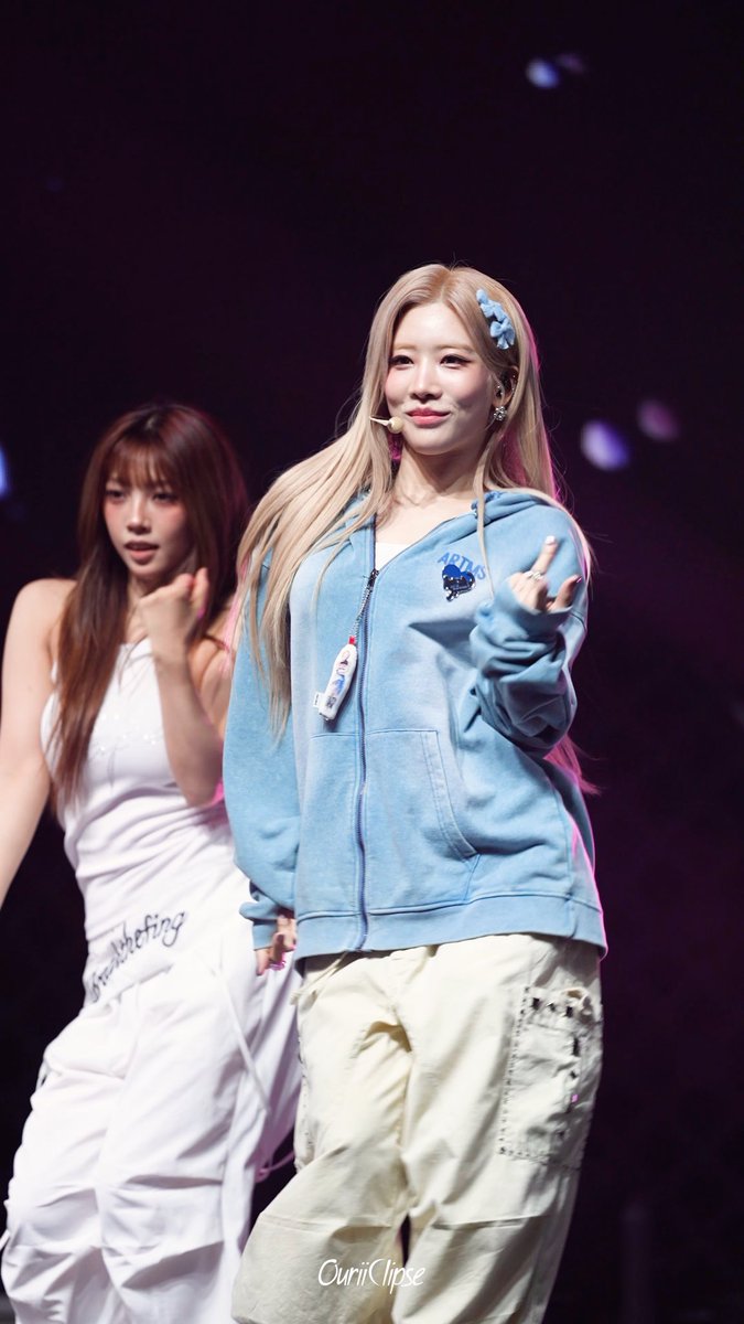 A concert photo of a blonde K-pop performer wearing a light blue hoodie with 'ABYSS' logo and cargo pants, smiling and making a hand gesture, with a backup dancer in white behind her; watermark 'OuriiOipse'.