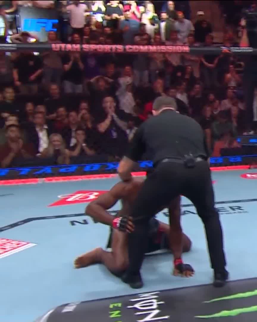 A UFC MMA fight inside an octagon with a referee in black separating two fighters on the mat; a crowd cheers in the background and 'UTAH SPORTS COMMISSION' and 'UFC' signage is visible on the cage.