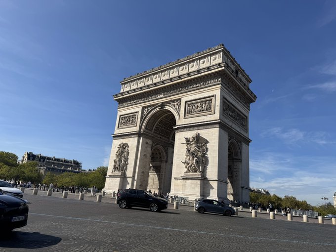 The Arc de Triomphe stands majestically under a clear blue sky, with cars including a black SUV and a "FR-B25" license-plated vehicle visible on the surrounding cobblestone streets. The image aligns with the post about being in Paris, as the Arc de Triomphe is one of the city's most iconic landmarks—suggesting the author is in the French capital balancing sightseeing with following Paris Fashion Week (PFW).