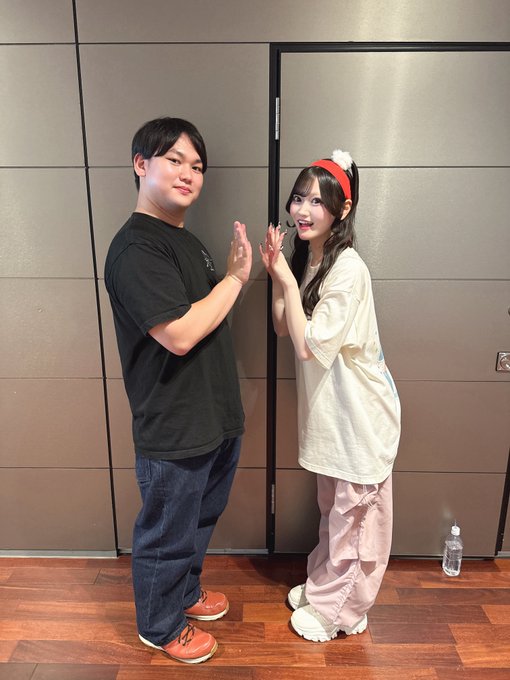 Two people stand facing each other with hands pressed together in a praying gesture in front of a paneled wall, with the person on the right wearing a red headband and oversized beige shirt.