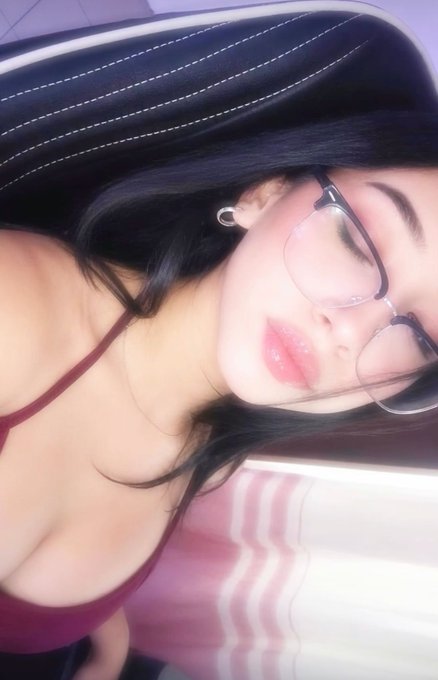 A close-up selfie of a young woman wearing glasses, a dark hat with white stitching, and a maroon top, lying down with her eyes closed.