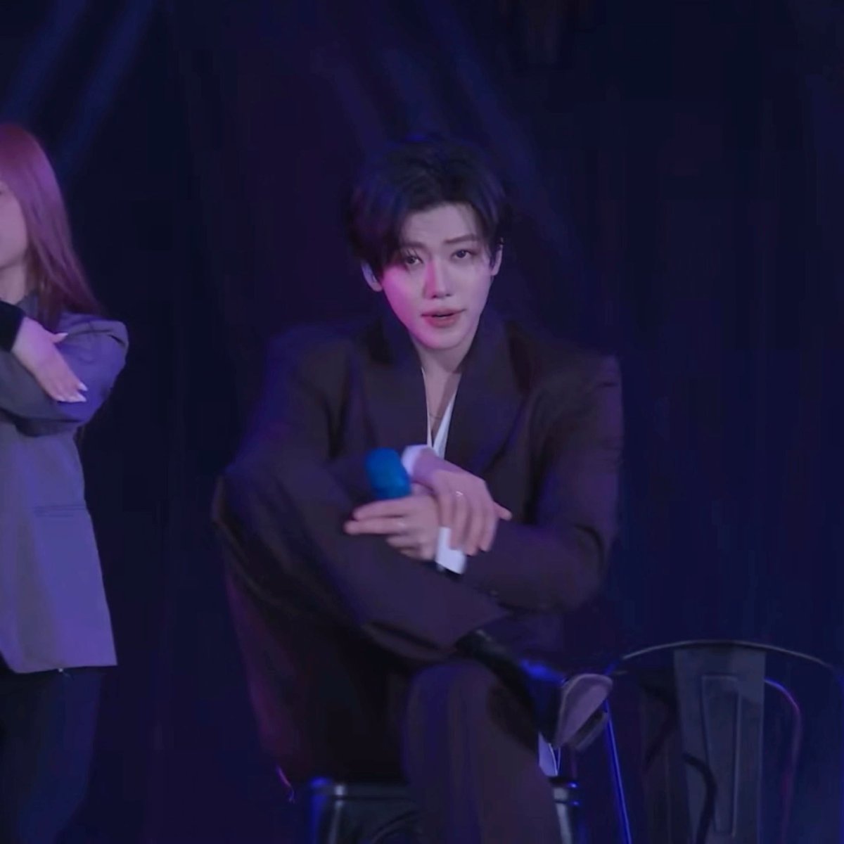 A young man in a dark brown suit holds a blue microphone while seated on stage in dim purple lighting.